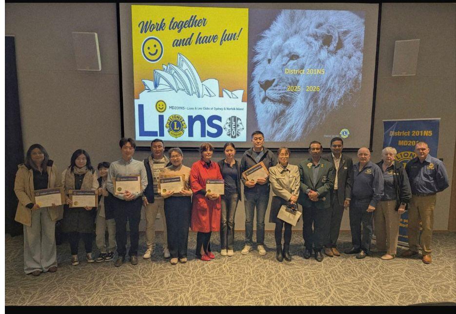SMULC members holding their Lions membership certificates at the District 201N5 charter presentation, with a 'Work together and have fun' District screen behind them