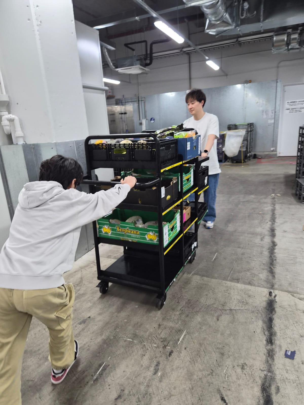 SMULC volunteers wheeling a trolley stacked with crates of rescued fruit and vegetables through an Aldi loading dock during a weekly food rescue shift